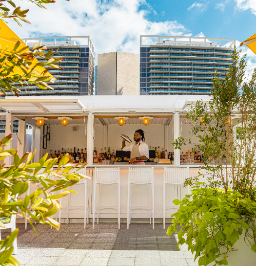 Outdoor bar area with bartender, white stools, plants, and modern city skyline in background.