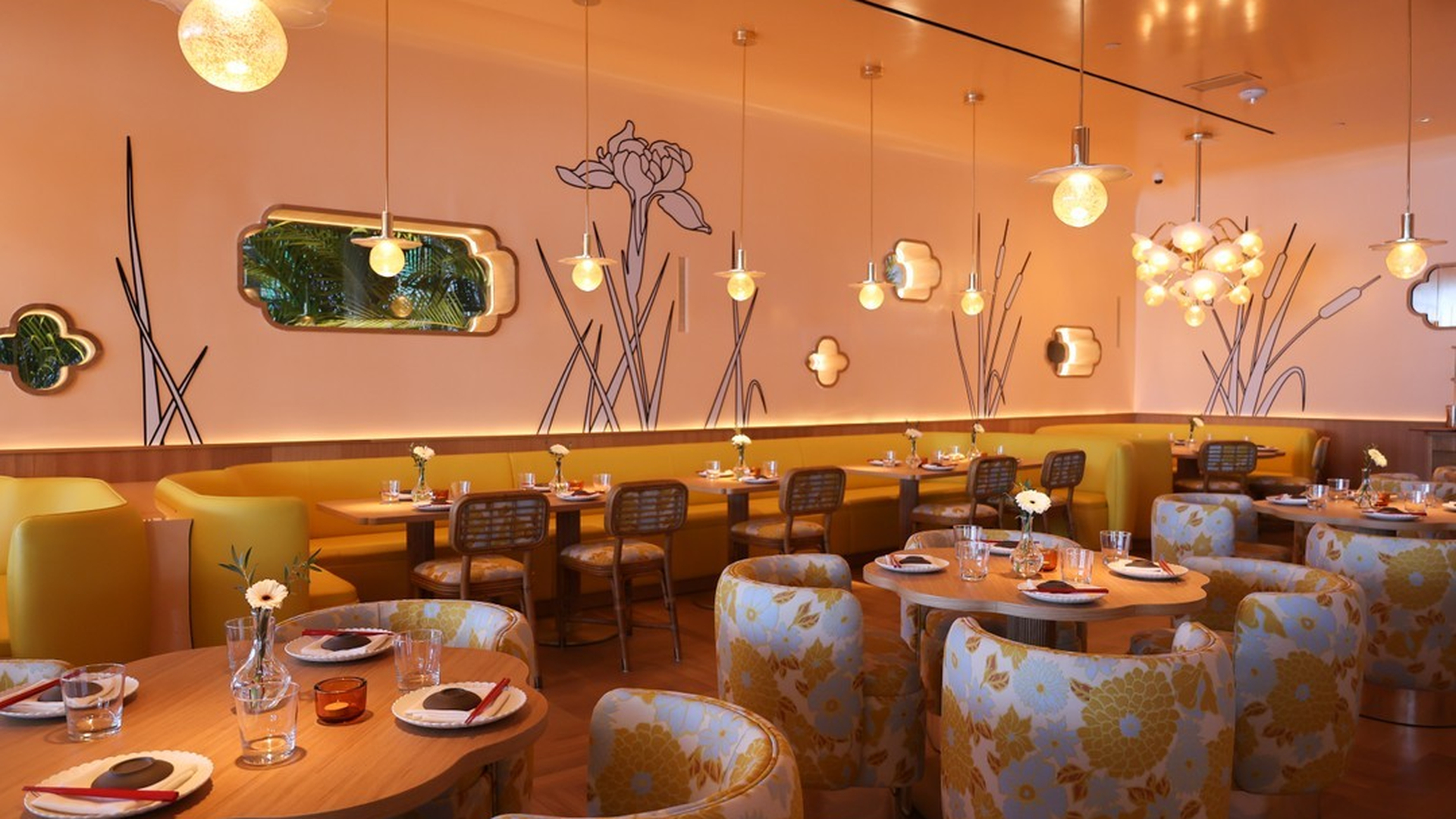 Restaurant interior with yellow walls, floral wall art, patterned chairs, and round tables set for dining.